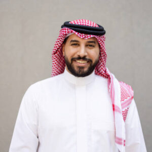 1 (1) Portrait of a happy young Saudi businessman standing against a gray wall and looking at the camera with a smile. Bearded Arab man wearing a thawb, kaffiyeh, and Agal.